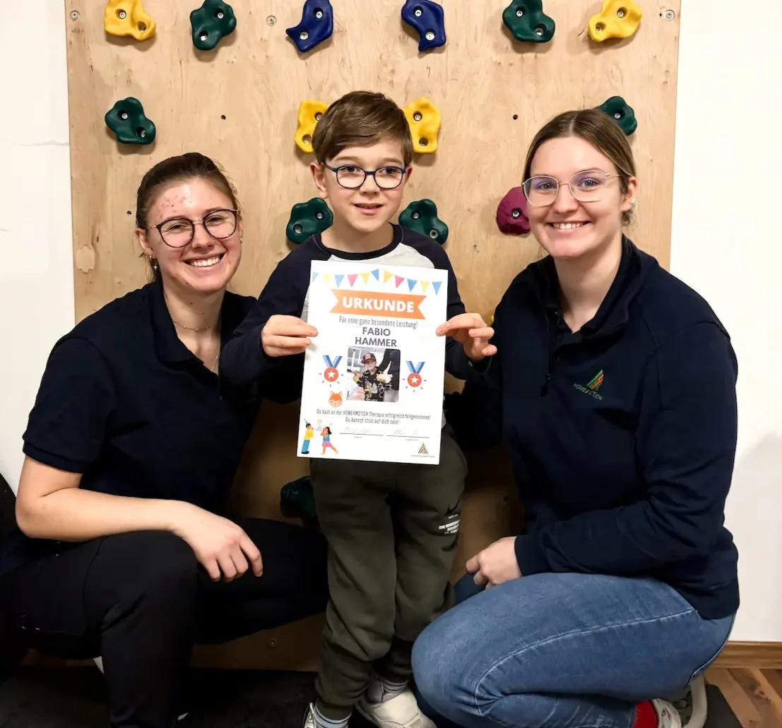 A child holding a certificate between two therapists in front of a climbing wall