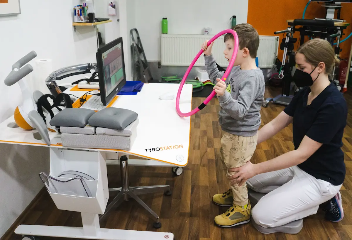 A child reaches for a hoop while a therapist supports the leg; a therapy device with a screen alongside