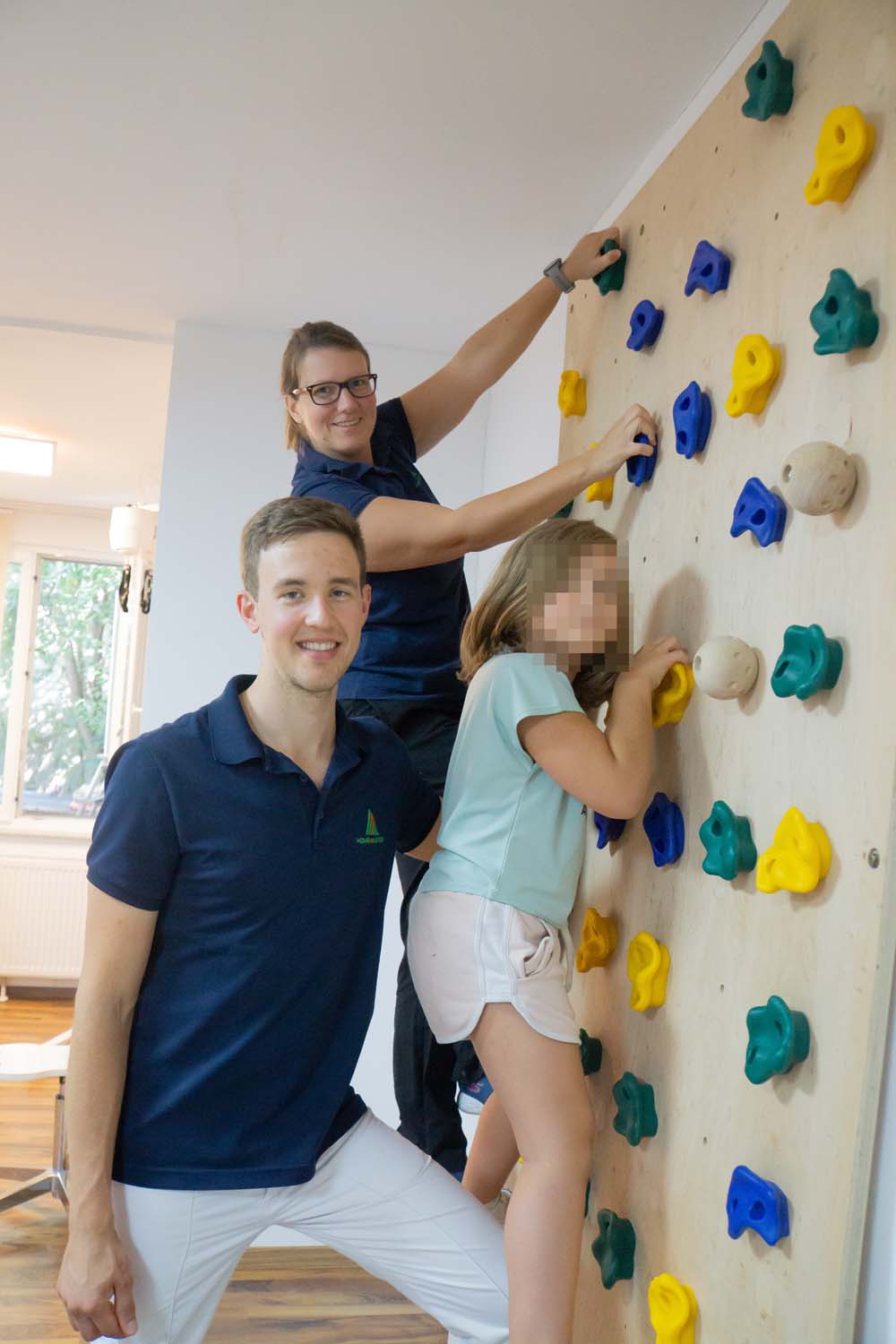 Lara auf einer Kletterwand mit ihren Physiotherapeut:innen Martina & Lorenz