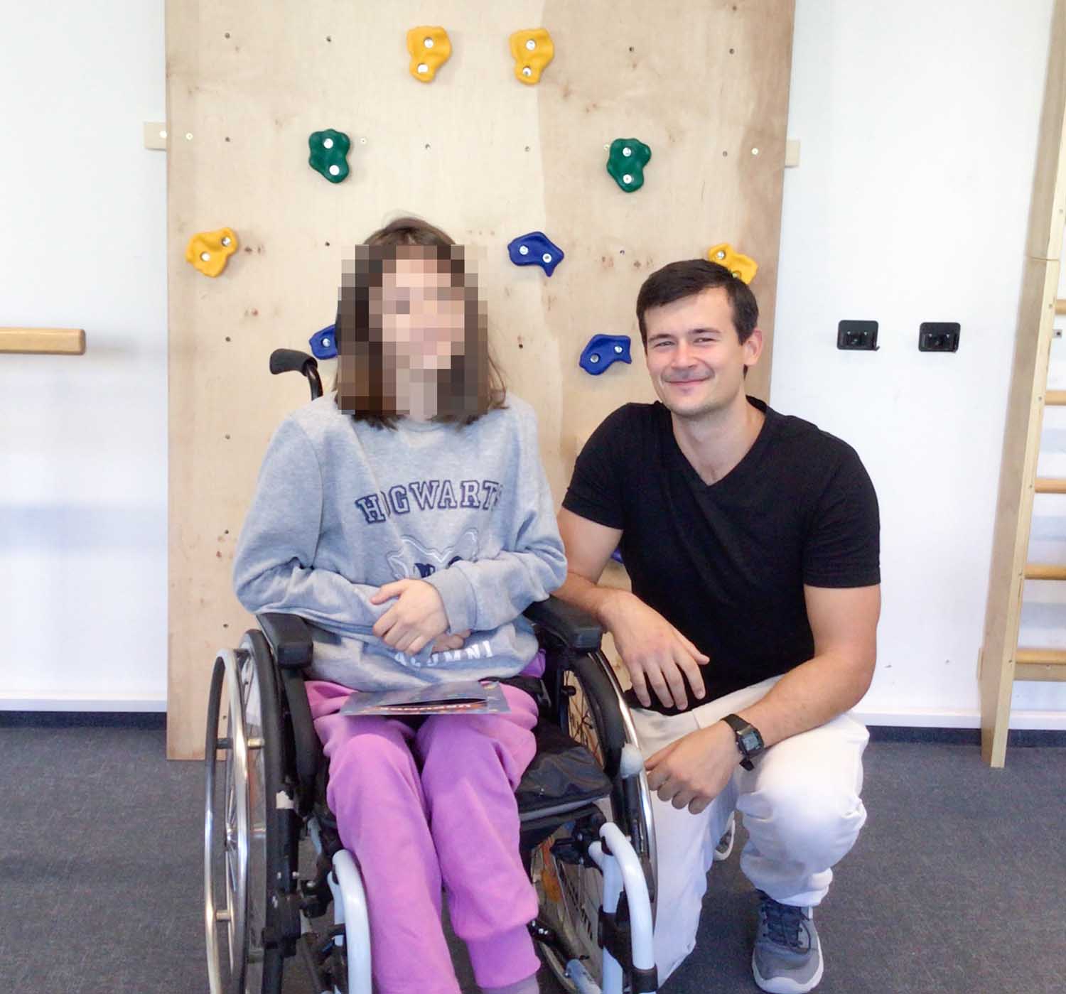 Lejla im Rollstuhl mit rosa Hose und grauem Tshirt. im Hintergrund eine Kletterwand. Therapeut Constantin kniet neben ihr
