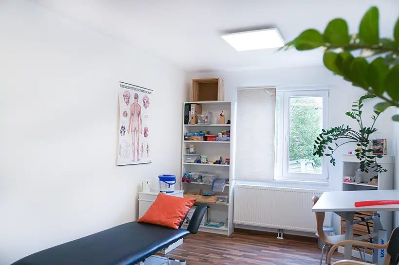Behandlungsraum mit Therapieliege, Regal mit Therapiematerial und einer Anatomietafel