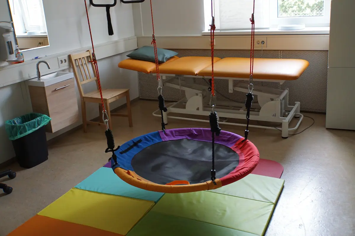 Nestschaukel über einer bunten Mattenfläche, daneben eine höhenverstellbare Therapieliege