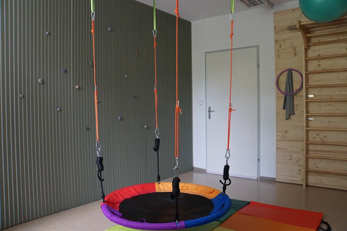 Nestschaukel an bunten Seilen über gelb-grünen Bodenmatten, an der Wand eine Sprossenwand aus Holz