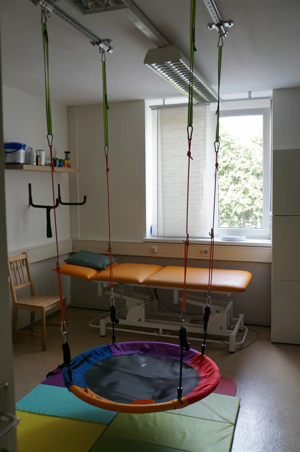 Therapieraum in Graz mit bunter Nestschaukel über einer Weichbodenmatte, dahinter eine Therapieliege am Fenster