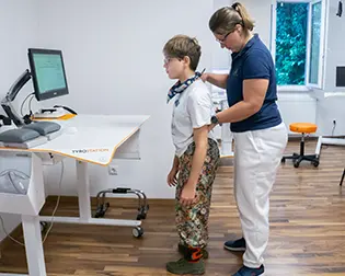 A therapist supports a child standing at a therapy device with a screen
