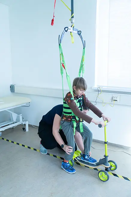 A child walks with a safety harness on a ceiling rail system, using a walking frame